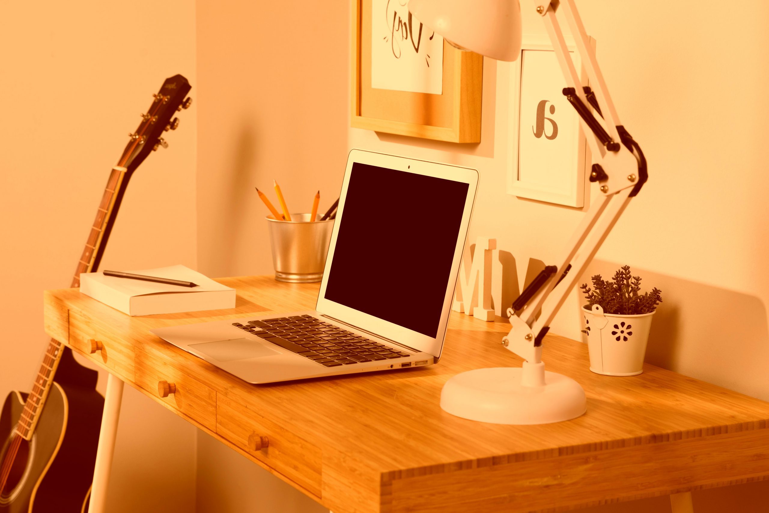 Escritorio con portátil , flexo y material de oficina, con guitarra al fondo apoyada en la mesa. Imagen de tonos anaranjados.