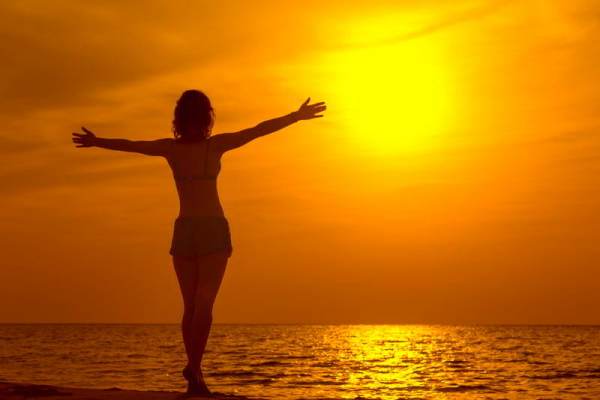 Mujer con los brazos abiertos frente al sol en una playa al atardecer. Importancia de la iluminación natural en tu espacio de trabajo.