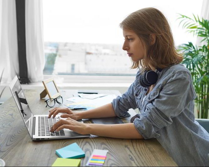 Mujer escribiendo en un portátil en un despacho bien iluminado por una gran ventana. Cómo iluminar un espacio de trabajo.