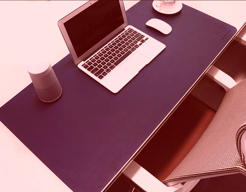 Alfombrilla grande azul sobre escritorio, con un portátil encima y un ratón, al lado se ve parte de una silla de oficina.